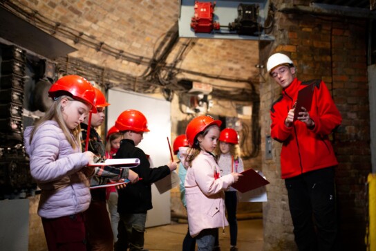 Grupa dzieci w czerwonych kaskach i z podkładkami do notowania patrzy na przewodnika w białym kasku.