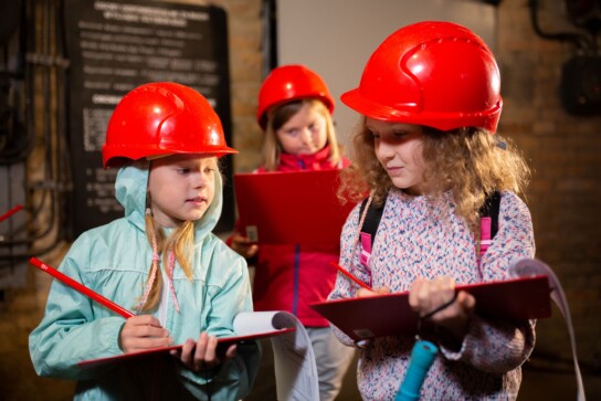 Dwie dziewczynki w czerwonych kaskach na głowie notują ołówkiem na trzymanych przez siebie podkładkach