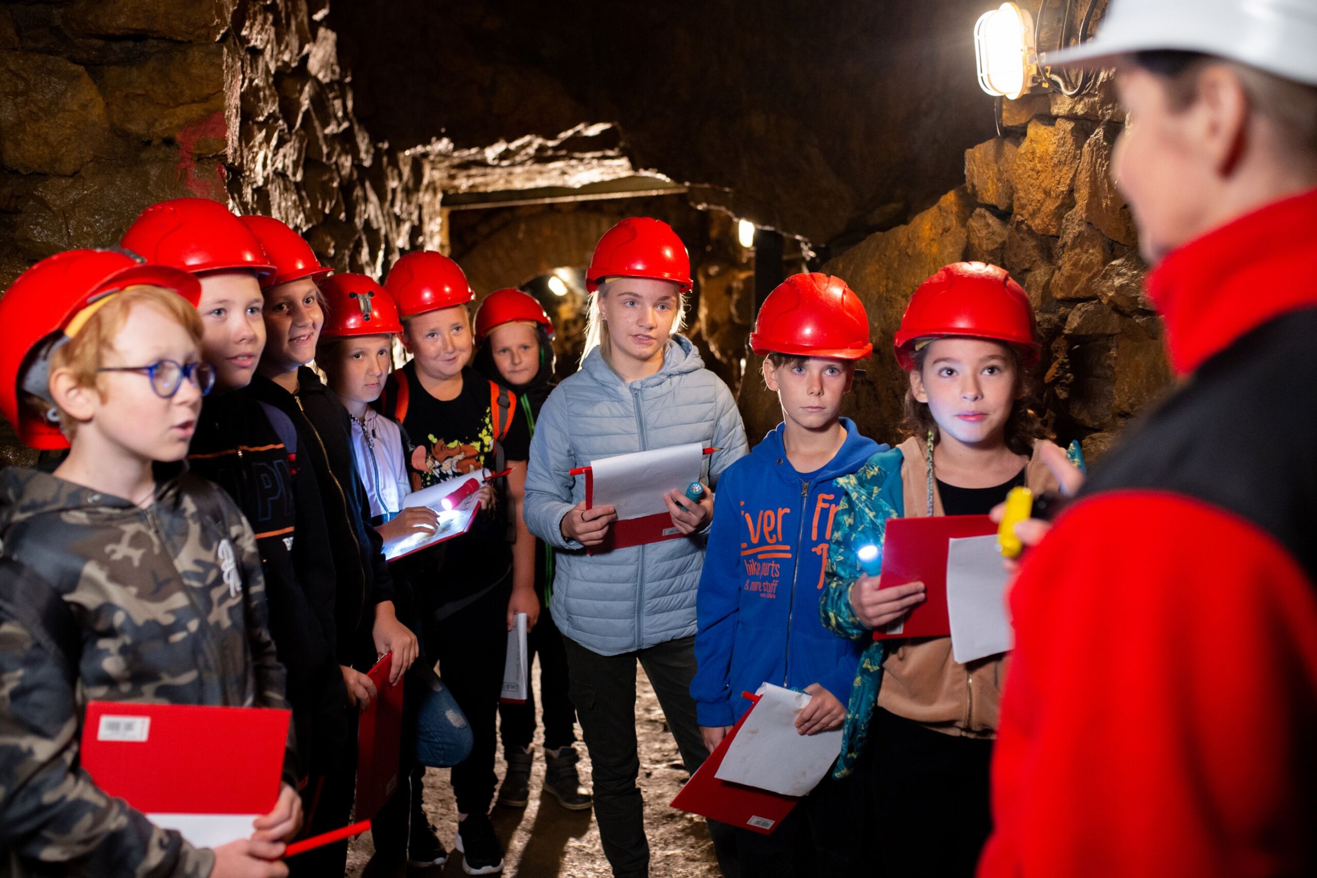 Grupa dzieci w czerwonych kaskach i z podkładkami do notowania patrzy na przewodniczkę w białym kasku.