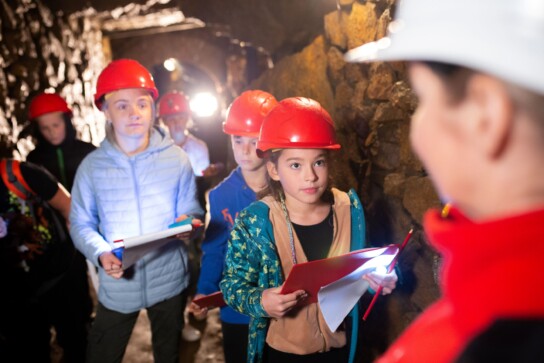 Grupa dzieci w czerwonych kaskach i z podkładkami do notowania patrzy na przewodniczkę w białym kasku.